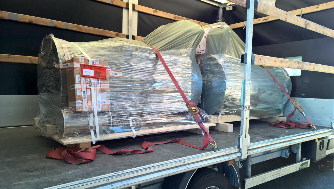 Ładunek zapakowany w folię stretch i zabezpieczony pasami transportowymi na platformie ciężarówki, częściowo przykryty zieloną plandeką.