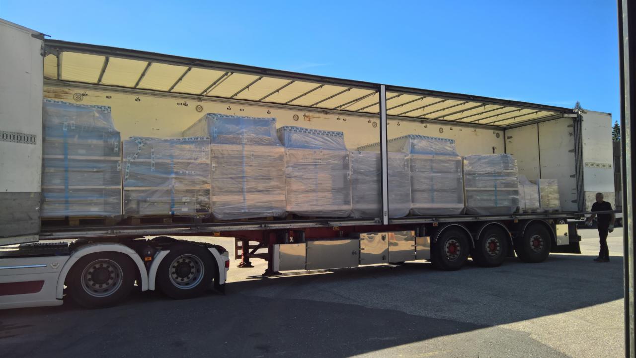 Otwarta naczepa ciężarówki z ładunkiem palet owiniętych folią stretch, widoczny fragment kabiny i opony, na zewnątrz stoi mężczyzna.