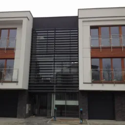 Nowoczesna fasada budynku z drewnianymi oknami i balkonami z balustradami ze stali nierdzewnej. Centralna część budynku przeszklona z poziomymi żaluzjami.