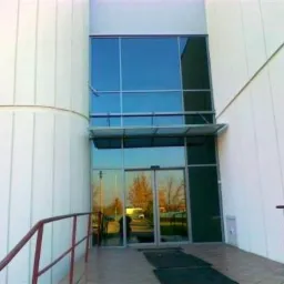 Szklane drzwi wejściowe z dużymi oknami w nowoczesnym budynku o białej fasadzie. Widoczne odbicie drzew w szybach.