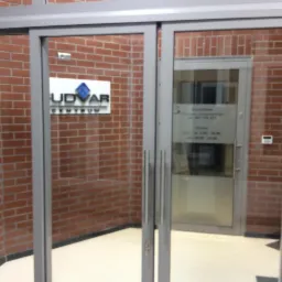 Szklane drzwi przesuwne z metalowymi ramami, widoczne na tle ceglanej ściany z logo firmy U dvar Centrum i godzinami otwarcia na szybie drzwi.