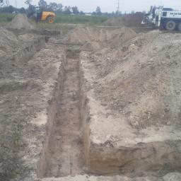 Ecodomus Przedsiębiorstwo Wielobranżowe - Wykopane wąskie rowy pod fundamenty z hałdami ziemi. W tle widoczny traktor i betoniarka. Prace ziemne w toku, teren budowy.