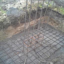 Ecodomus Przedsiębiorstwo Wielobranżowe - Zbrojenie fundamentu w wykopie, widok z góry. Stalowe pręty połączone w siatkę, wystające pionowe elementy. Surowy beton.