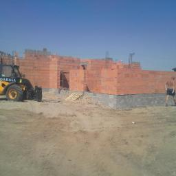 Ecodomus Przedsiębiorstwo Wielobranżowe - Budowa domu w toku: widoczne fundamenty z szarego betonu i ściany z czerwonej cegły. Na budowie stoi żółta ładowarka JCB, a obok przechadza się mężczyzna.