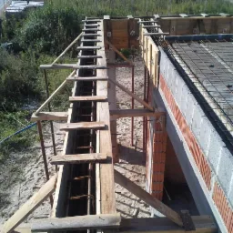 Zbrojenie fundamentów i szalunek z drewna. Widok z góry na plac budowy, z elementami konstrukcyjnymi i otoczeniem zieleni.