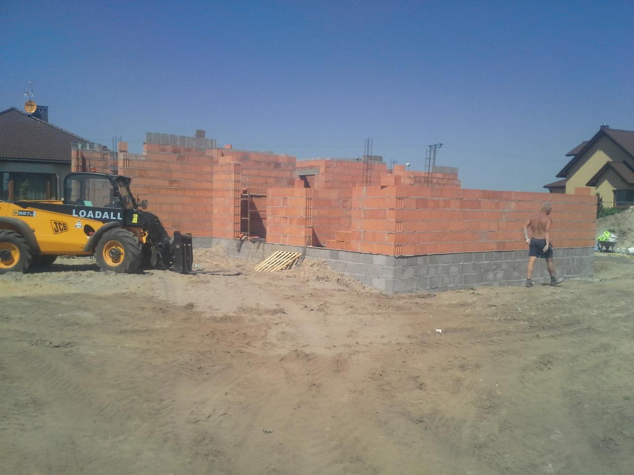Budowa domu w toku: widoczne fundamenty z szarego betonu i ściany z czerwonej cegły. Na budowie stoi żółta ładowarka JCB, a obok przechadza się mężczyzna.