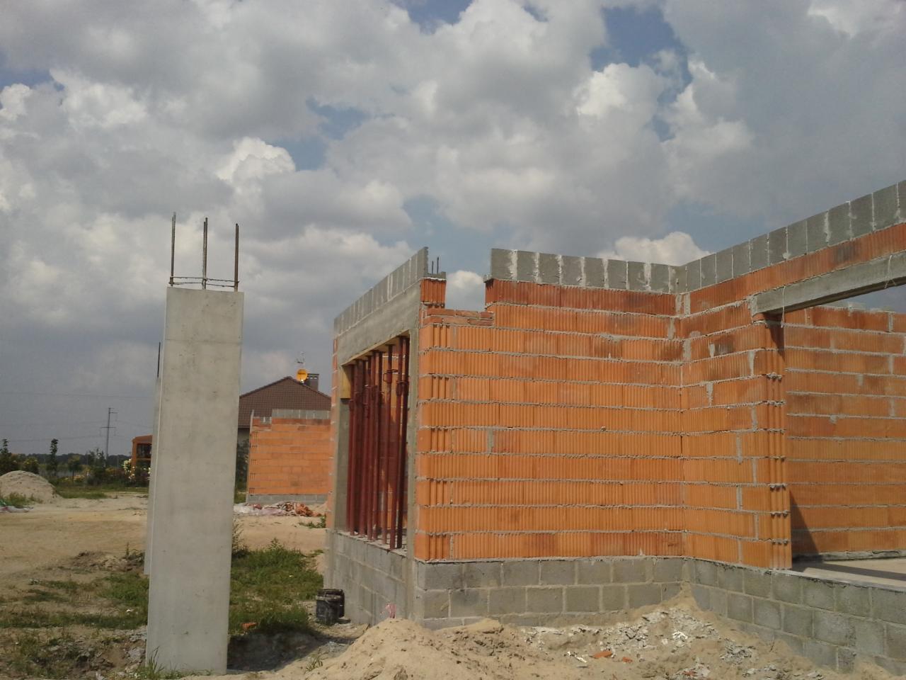 Wznoszenie ścian z czerwonej cegły na fundamencie z bloczków betonowych; widoczne zbrojenia słupów i stropu. Budowa domu w trakcie realizacji, błękitne niebo.