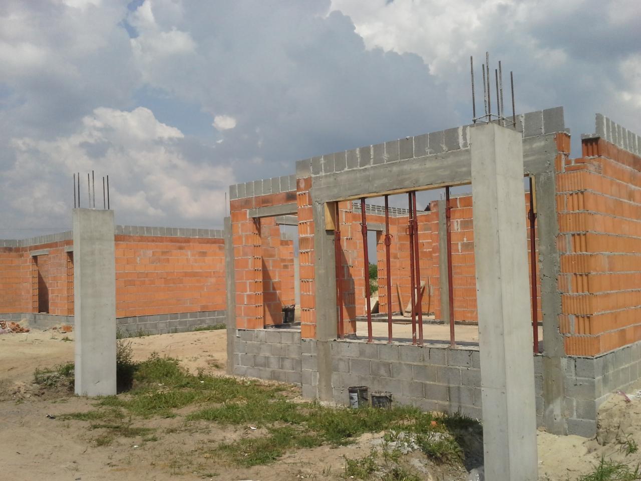 Budowa domu w trakcie wznoszenia ścian z czerwonej cegły i betonowych słupów, widoczne szalunki okienne i zbrojenia, słoneczny dzień.
