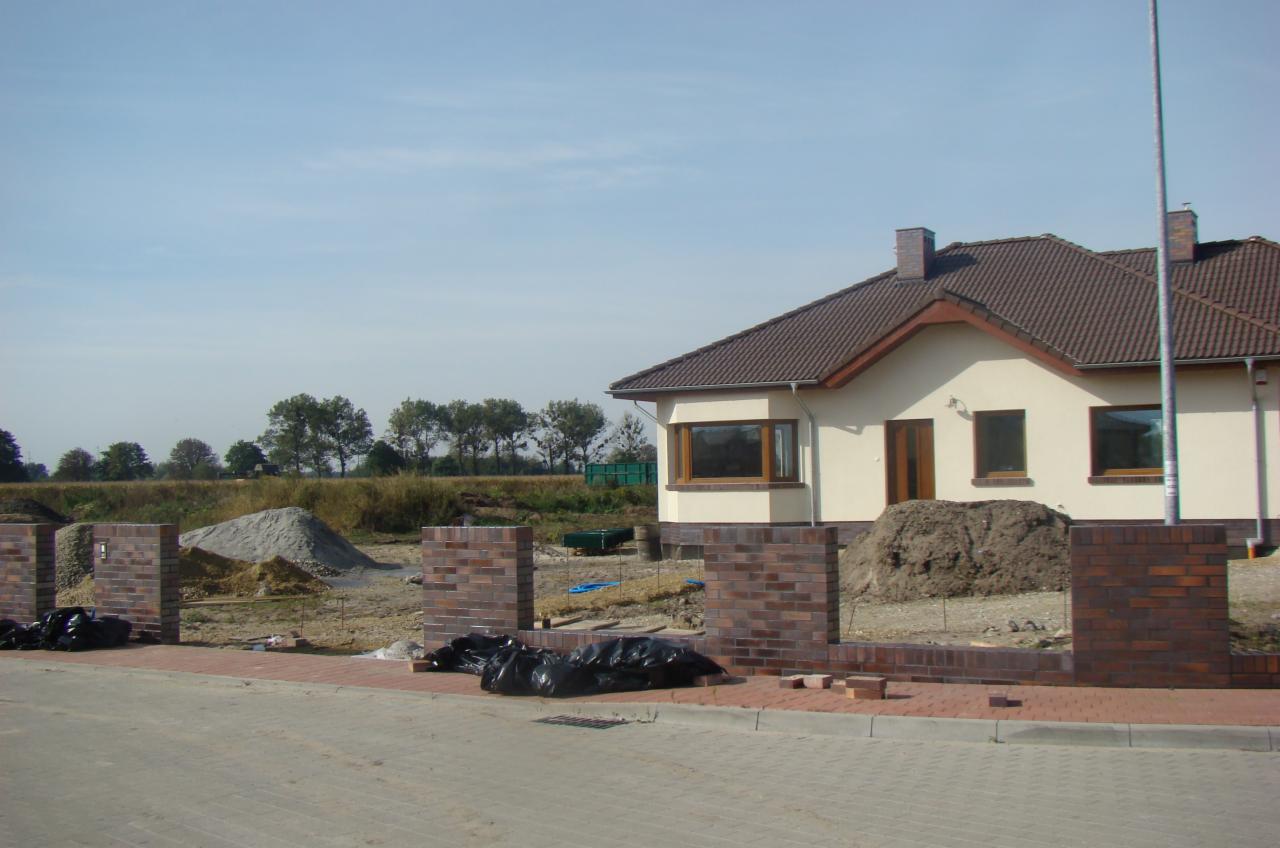 Dom z elewacją w trakcie budowy. Widoczne fragmenty muru z cegły klinkierowej, stosy piasku i ziemi. W tle drzewa i błękitne niebo.