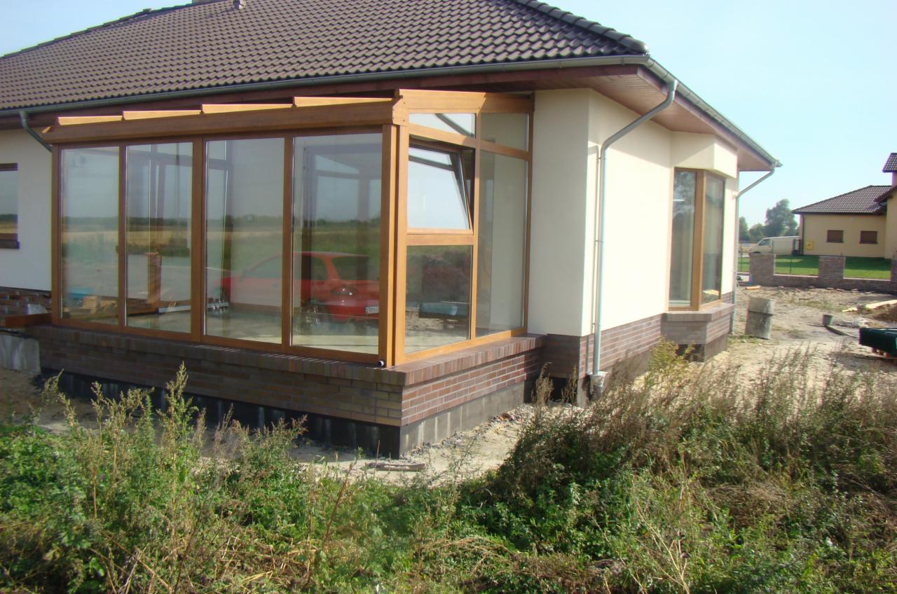 Jasna elewacja domu z dobudowaną, przeszkloną werandą na ceglanej podmurówce. Widok na otwarte okno i częściowo widoczny czerwony samochód wewnątrz.