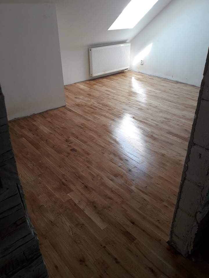 Jasne, drewniane panele podłogowe w pokoju na poddaszu z oknem dachowym, widoczny grzejnik i fragment surowej ściany.