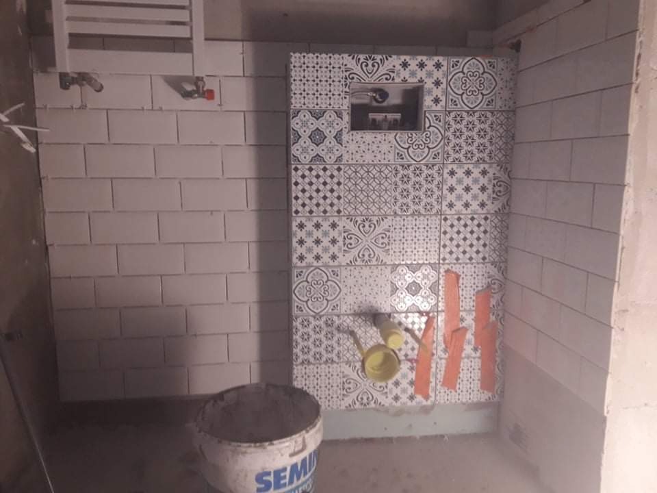 Układanie dekoracyjnych płytek w łazience, widoczny stelaż podtynkowy z otworem inspekcyjnym, białe płytki na ścianie obok i wiadro z zaprawą.