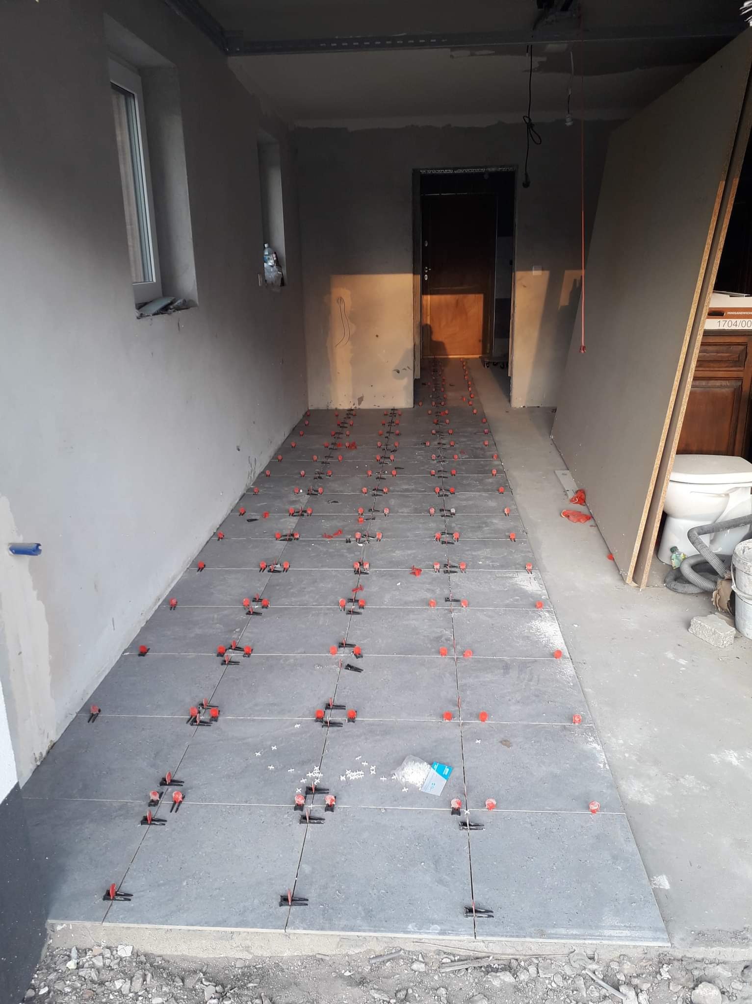 Szare płytki podłogowe układane w korytarzu z użyciem czerwonych klipsów poziomujących, widoczny fragment płyty karton-gips opartej o ścianę, surowy stan wykończenia.