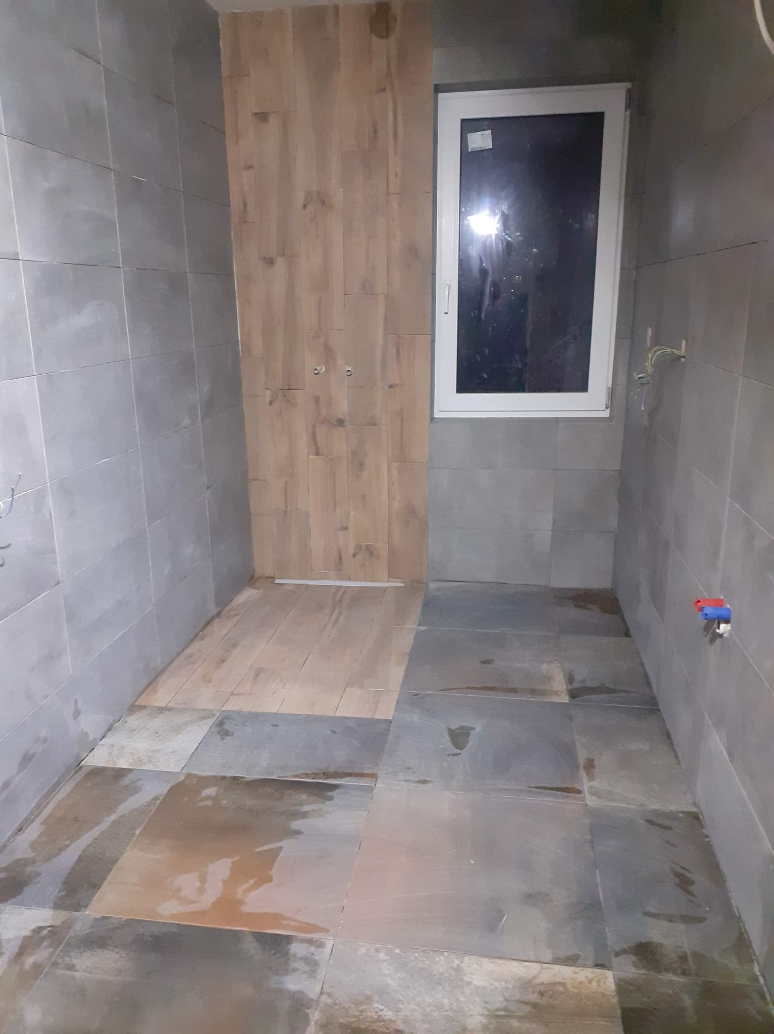 Wnętrze łazienki w trakcie remontu, z szarymi, kwadratowymi płytkami na ścianach i podłodze, drewnopodobnymi panelami na fragmencie ściany, białym oknem i wystającymi przewodami elektrycznymi.