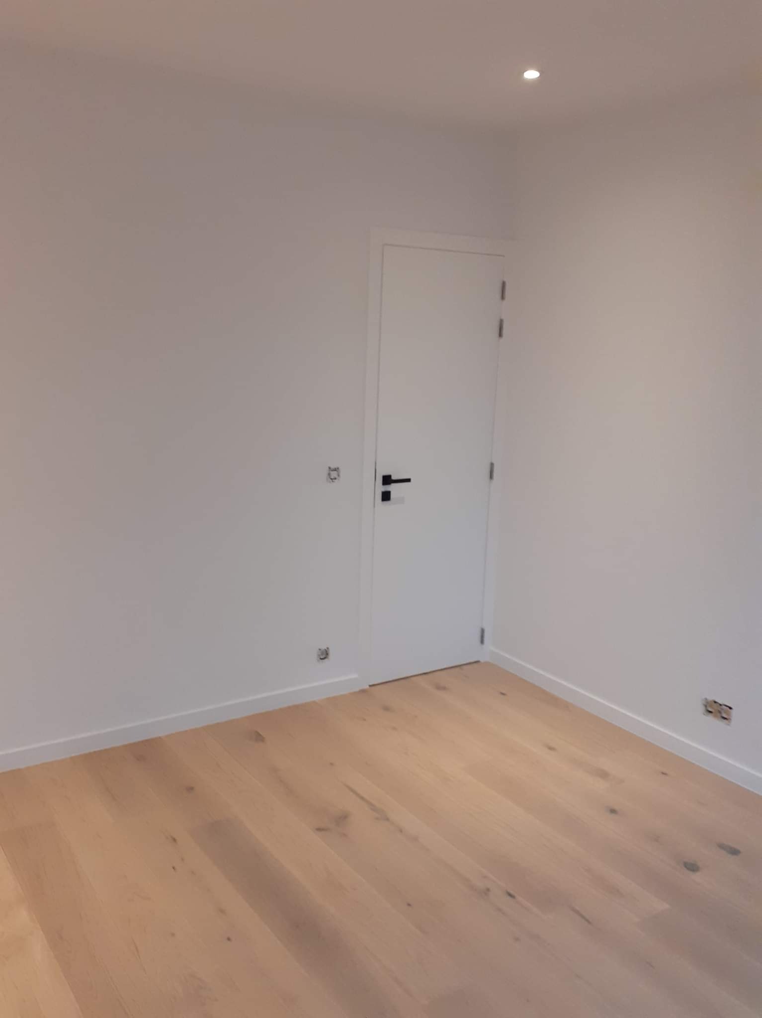 Pusty pokój z drewnianą podłogą, świeżo pomalowanymi białymi ścianami i białymi listwami przypodłogowymi. Widoczna biała, zamknięta, minimalistyczna  drzwi z czarną klamką. Na ścianie widoczne...