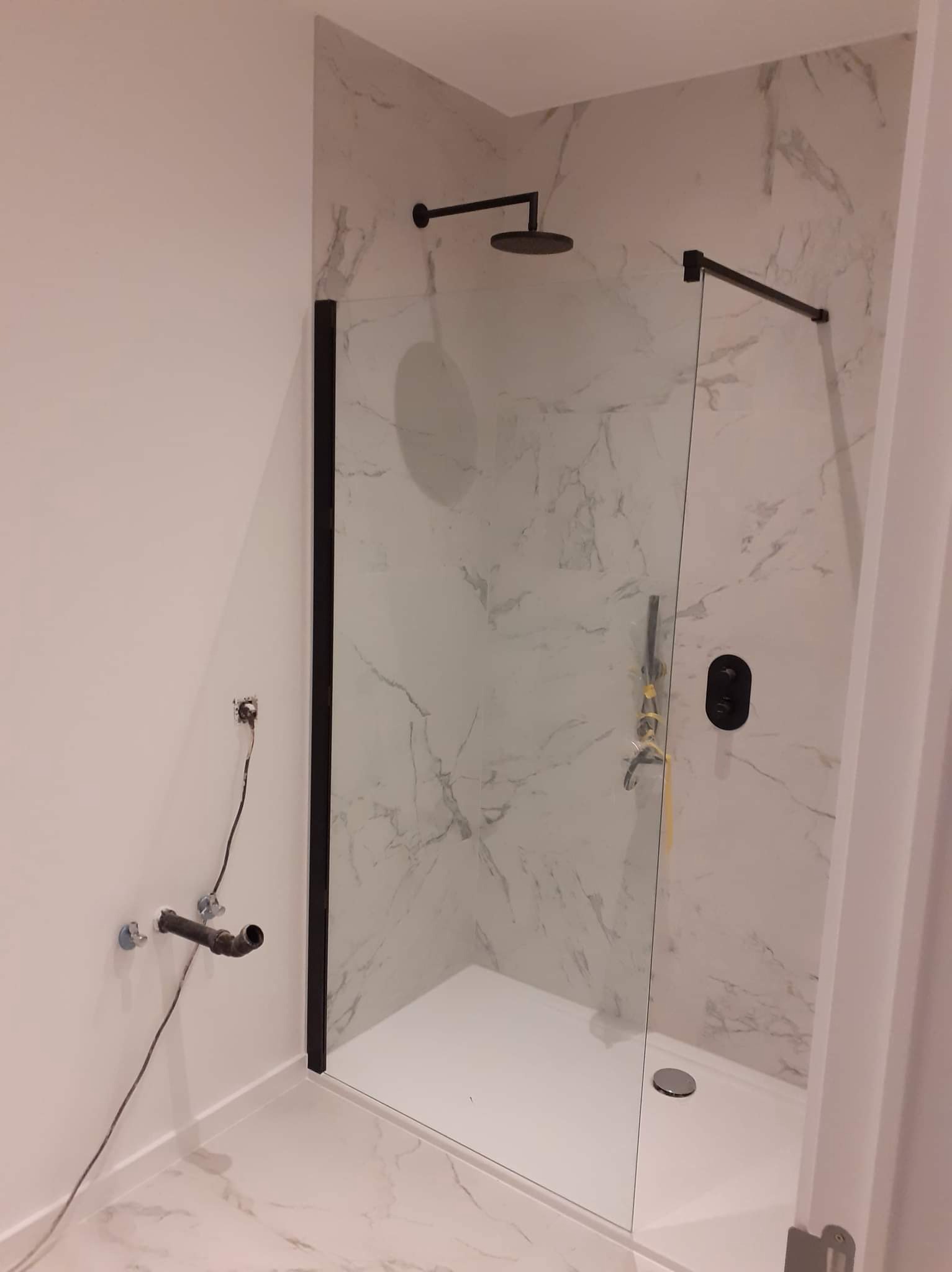 Nowoczesna kabina prysznicowa z czarnymi elementami i marmurowym wykończeniem ścian, widoczne niedokończone podłączenia hydrauliczne i elektryczne.
