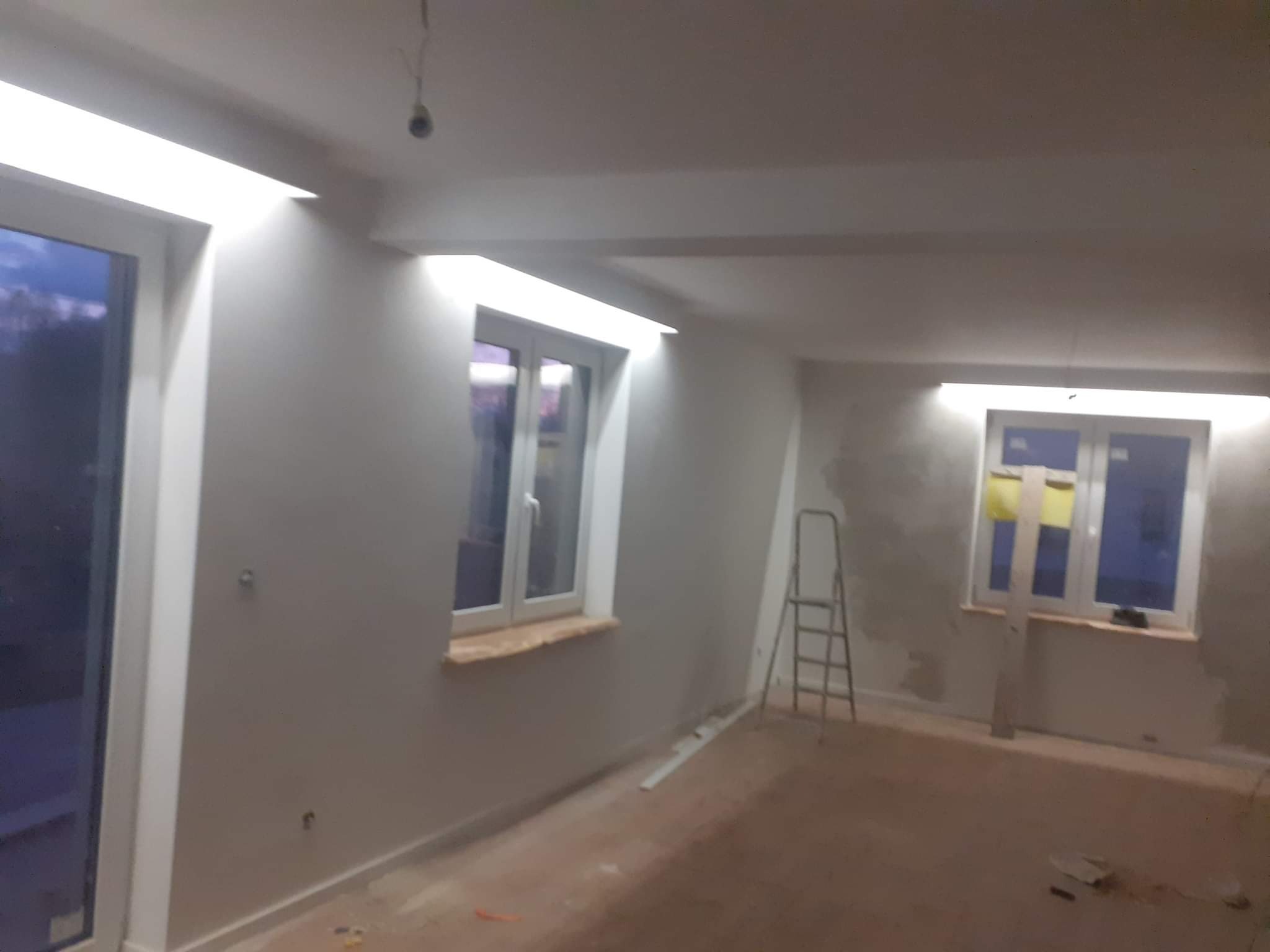 Wnętrze w trakcie malowania: ściany w szarym odcieniu, trzy okna z widokiem na zewnątrz, drabina oparta o ścianę, sufit z widocznymi punktami świetlnymi.
