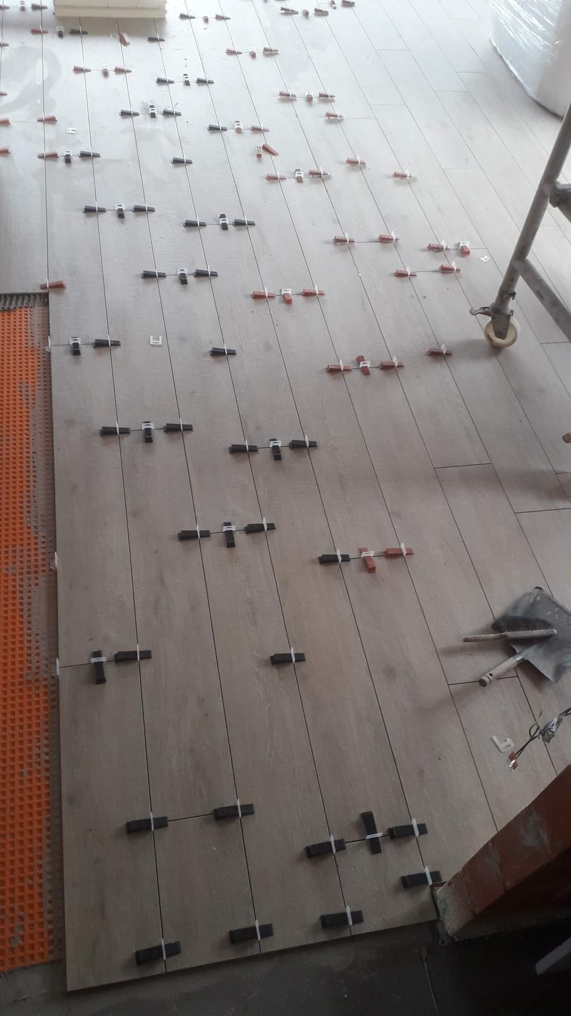 Ułożone płytki podłogowe imitujące drewno, z systemem poziomowania płytek (klipsy i kliny) oraz widoczną matą drenażową przy ścianie. W tle widoczny fragment rusztowania i narzędzia.