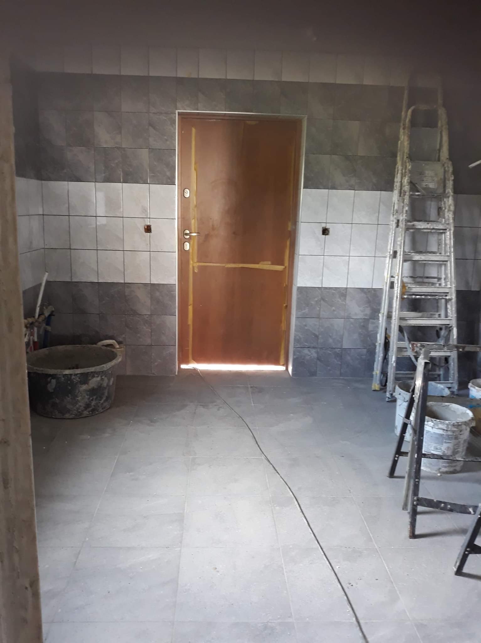 Wnętrze w trakcie remontu: ściany wyłożone szarymi i białymi płytkami, brudne wiadro i drabina oparta o ścianę, widoczne kable na podłodze.