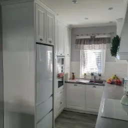 Jasna kuchnia z zabudową meblową w stylu klasycznym, z frontami frezowanymi, zintegrowaną lodówką i piekarnikiem, szarym blatem i okapem oraz oknem z roletami.