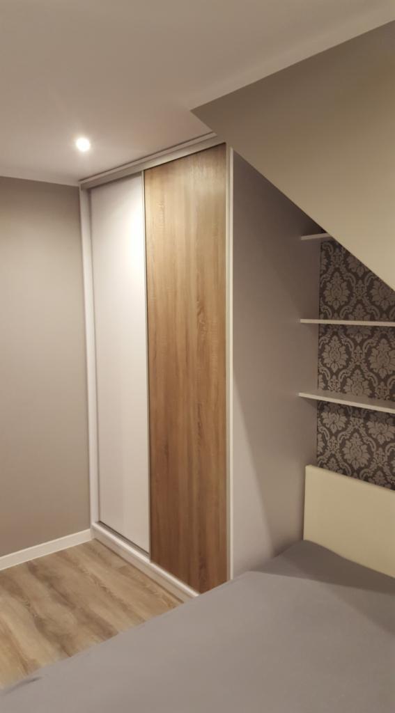Szafa przesuwna z dwoma drzwiami: jedno białe, drugie z okleiną imitującą jasne drewno, wbudowana we wnękę obok łóżka na tle ściany z tapetą w ornamenty.