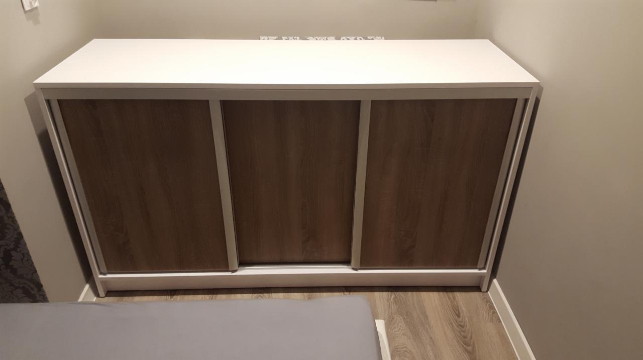 Zamknięta szafa wnękowa z przesuwnymi drzwiami o teksturze drewna i białym obramowaniu, widoczna częściowo z boku, na tle beżowej ściany i podłogi imitującej drewno.