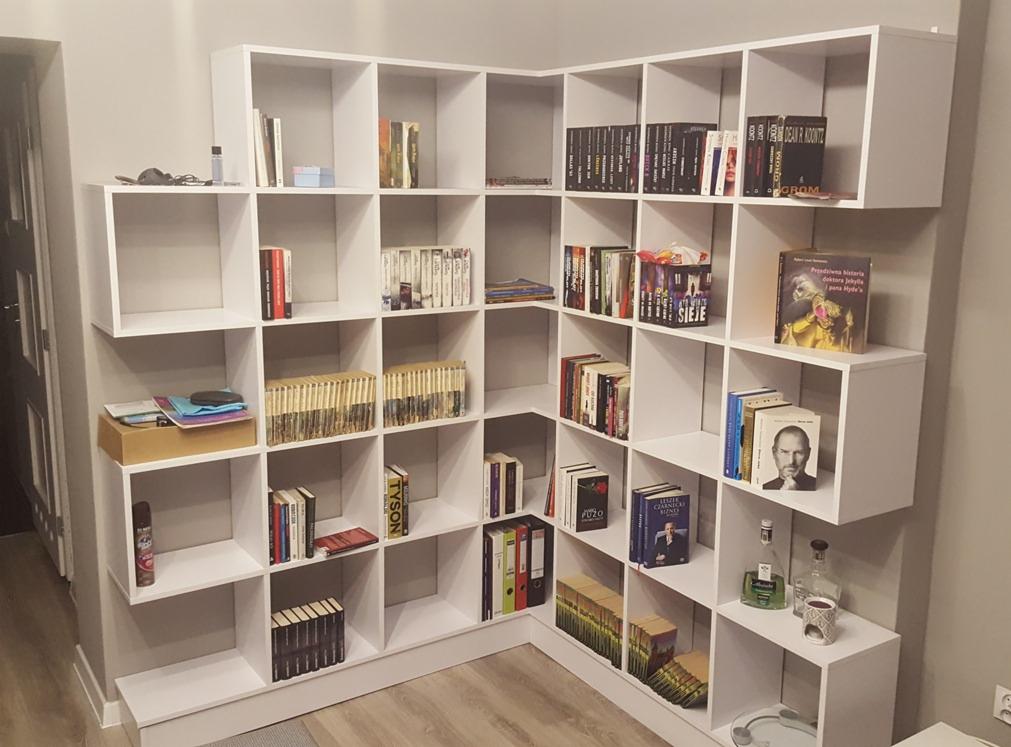 Biały, narożny regał na książki z półkami o kwadratowych modułach, wypełniony książkami, dekoracjami i akcesoriami biurowymi, stojący na podłodze z paneli.