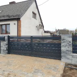 Nowoczesna, przesuwna brama wjazdowa w kolorze grafitowym z poziomymi panelami, osadzona na słupkach z szarej kostki imitującej kamień, przed domem z białą elewacją i szarym dachem.