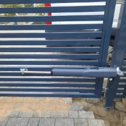 Szara, pozioma brama wjazdowa z widocznym siłownikiem automatyki i fragmentem zaparkowanego pojazdu w tle.