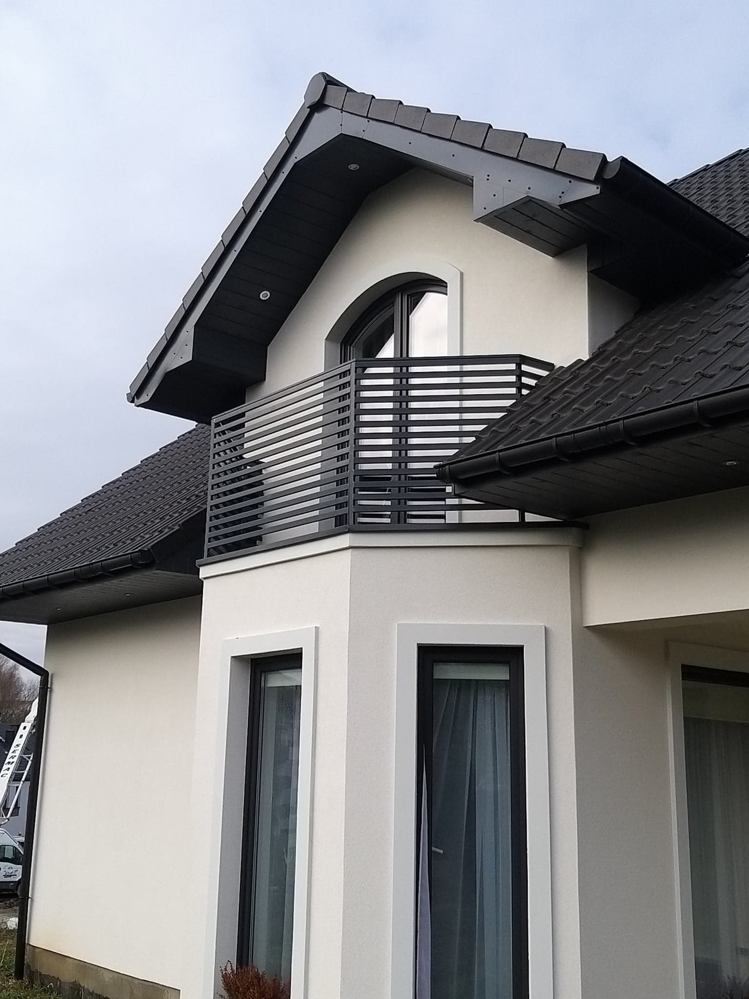 Elewacja domu z balkonem zabezpieczonym nowoczesną, poziomą, metalową balustradą w kolorze grafitowym, widok z dołu na fragment budynku z dwoma oknami.