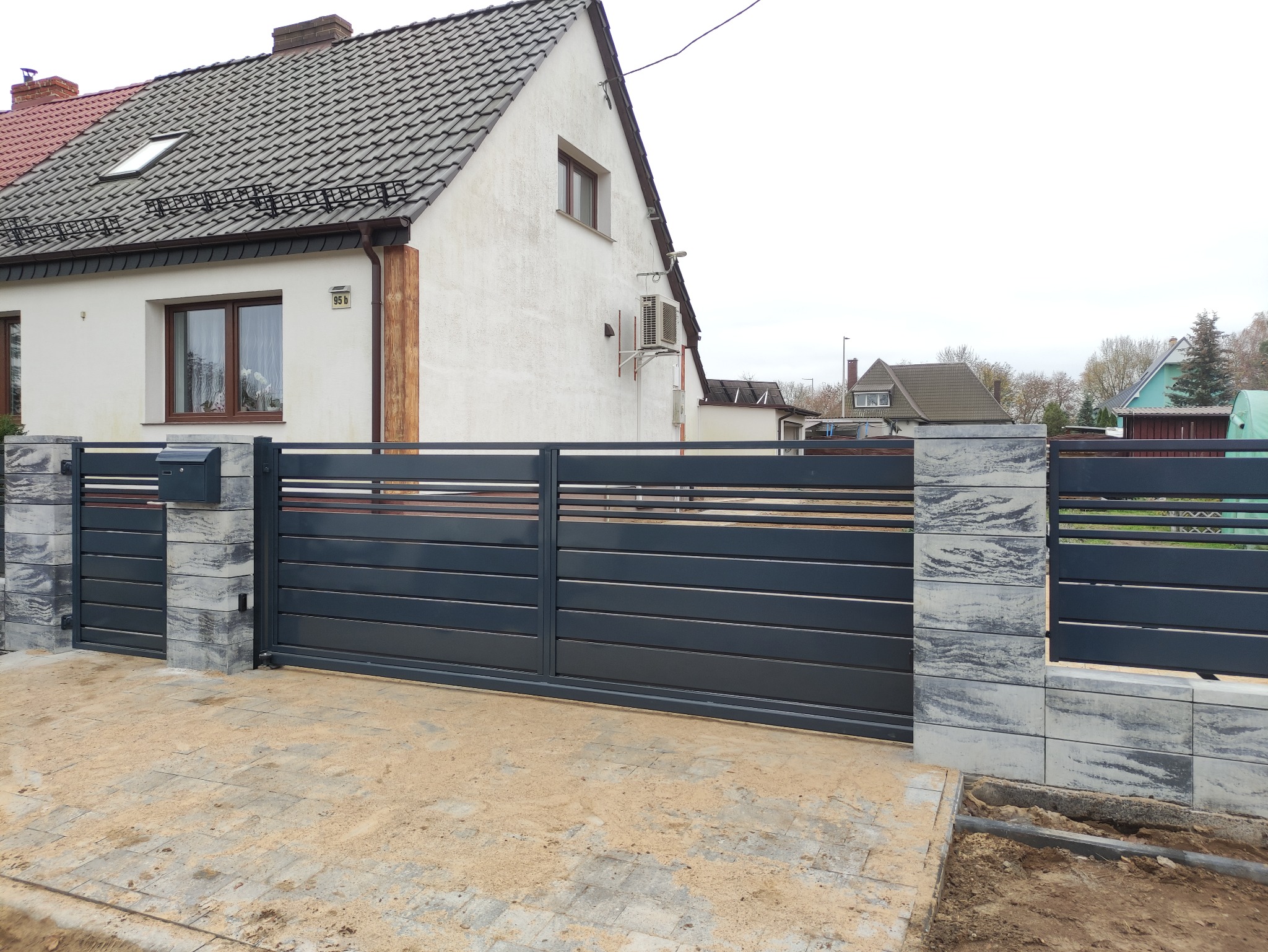 Nowoczesna, przesuwna brama wjazdowa w kolorze grafitowym z poziomymi panelami, osadzona na słupkach z szarej kostki imitującej kamień, przed domem z białą elewacją i szarym dachem.