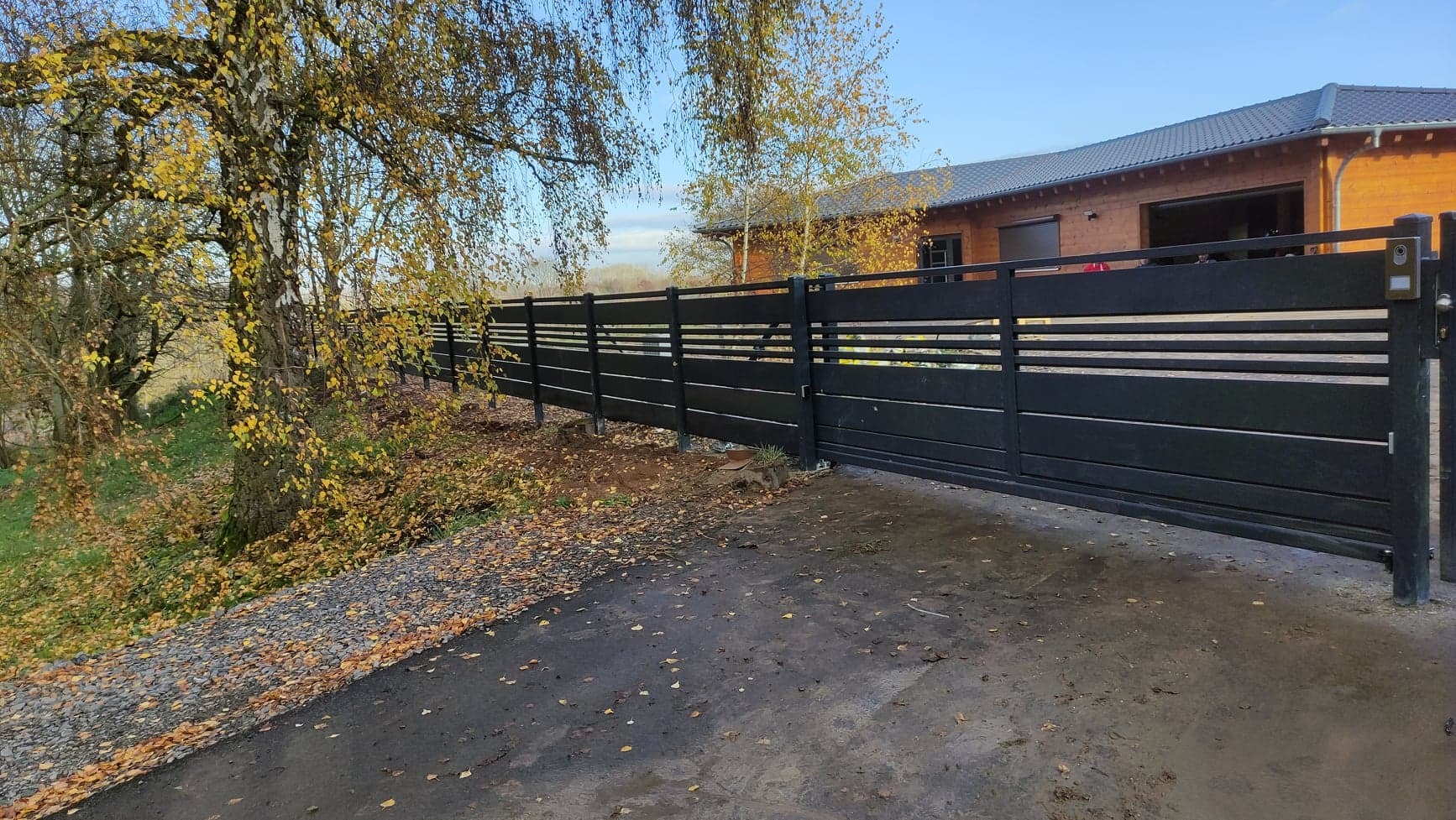 Nowoczesna, przesuwna brama wjazdowa z poziomymi panelami, zamontowana przy posesji z drewnianym domem i drzewami z żółtymi liśćmi w tle.