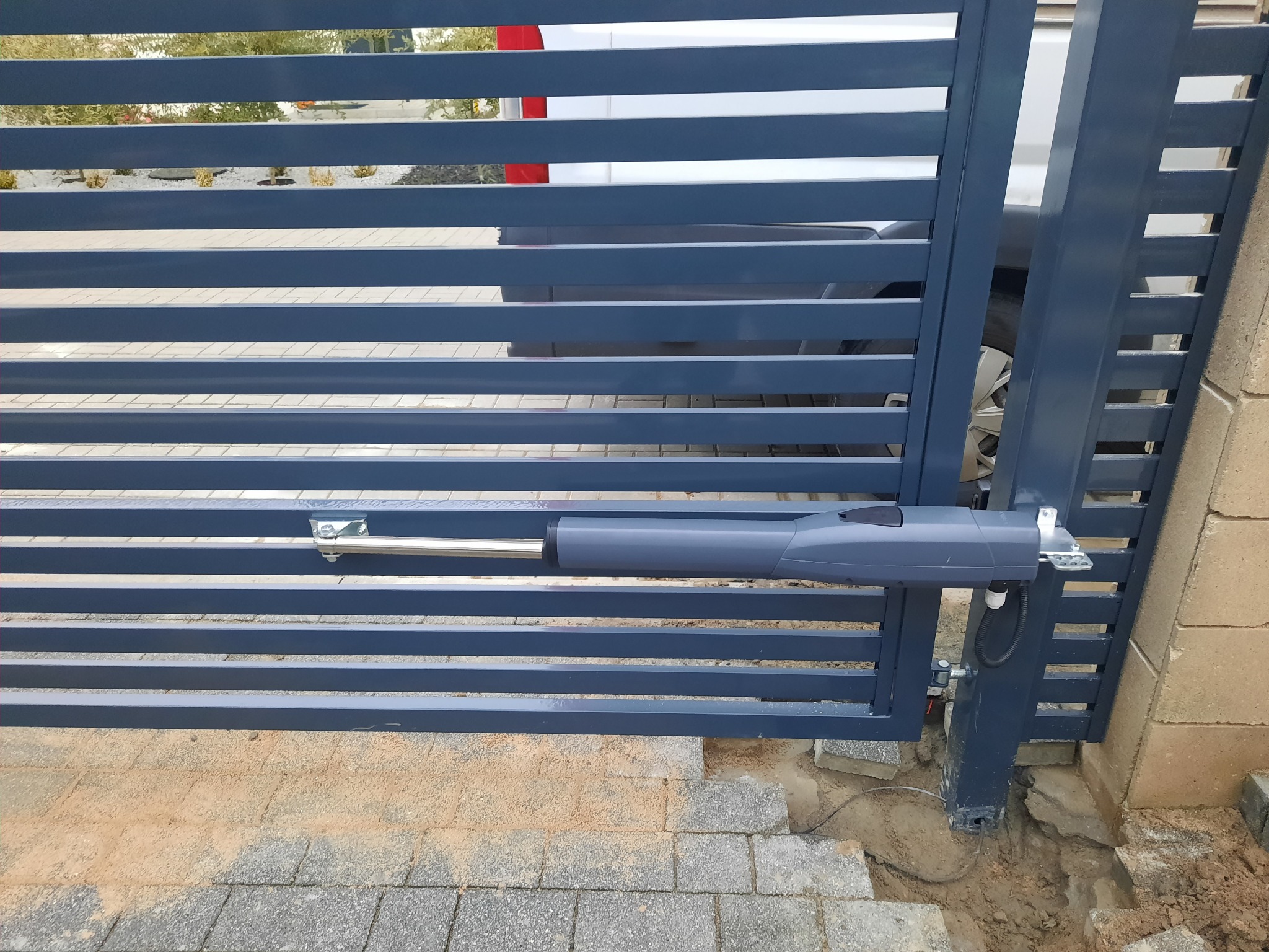 Szara, pozioma brama wjazdowa z widocznym siłownikiem automatyki i fragmentem zaparkowanego pojazdu w tle.