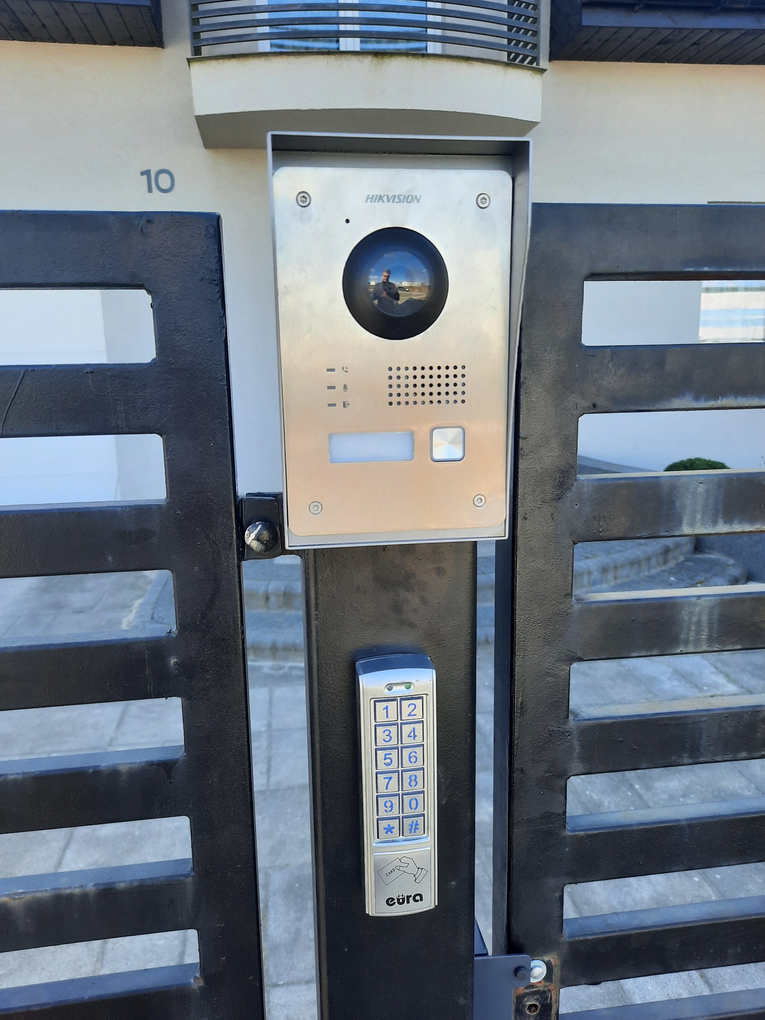 Czarna brama wjazdowa z domofonem Hikvision i klawiaturą numeryczną Eura, zamontowana przy ścianie budynku z numerem 10.