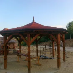 Drewniana altana z czerwonym dachem i piec chlebowy z kamienia w tle, na piaszczystym podłożu.