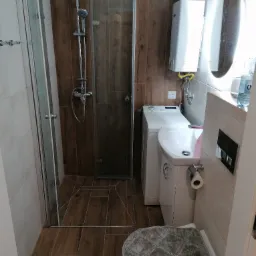 Ukończona łazienka z kabiną prysznicową, pralką zabudowaną obok toalety i umywalki, drewnopodobne płytki na podłodze i ścianie, podświetlane lustro owalne.