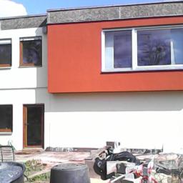 F.W-BUD Marcin Kruczyński - Dwukondygnacyjny dom z pomarańczową ścianą, widoczne wiadro, gruz i taśma ostrzegawcza sugerujące prace budowlane na zewnątrz.