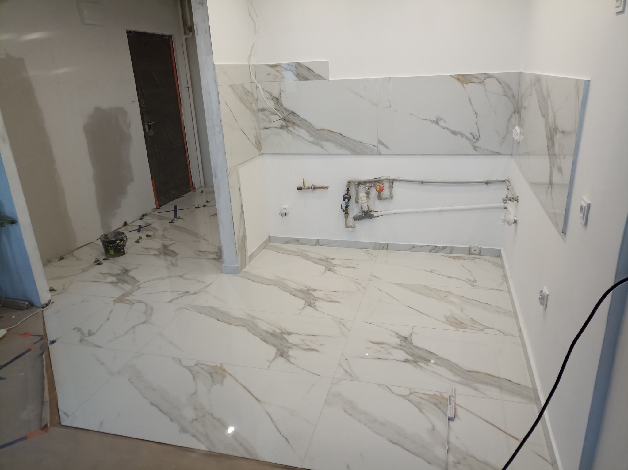 Białe płytki imitujące marmur na podłodze i ścianach łazienki w trakcie remontu w Rydułtowach, widoczne krzyżyki dystansowe i wiadro z klejem.