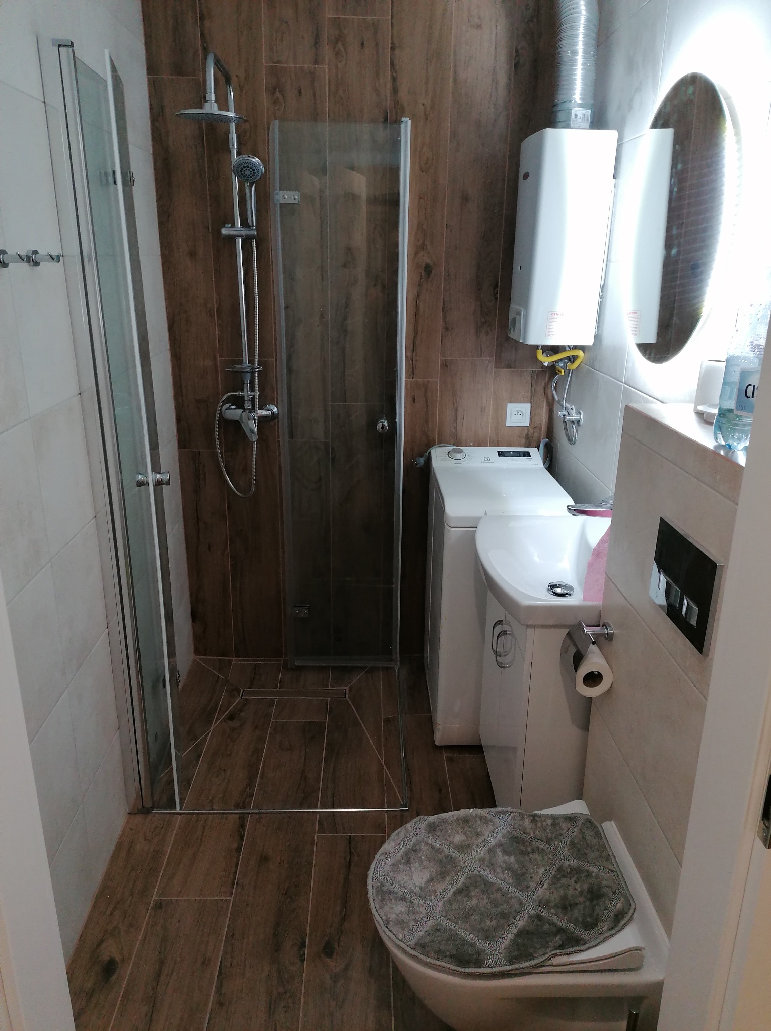 Ukończona łazienka z kabiną prysznicową, pralką zabudowaną obok toalety i umywalki, drewnopodobne płytki na podłodze i ścianie, podświetlane lustro owalne.