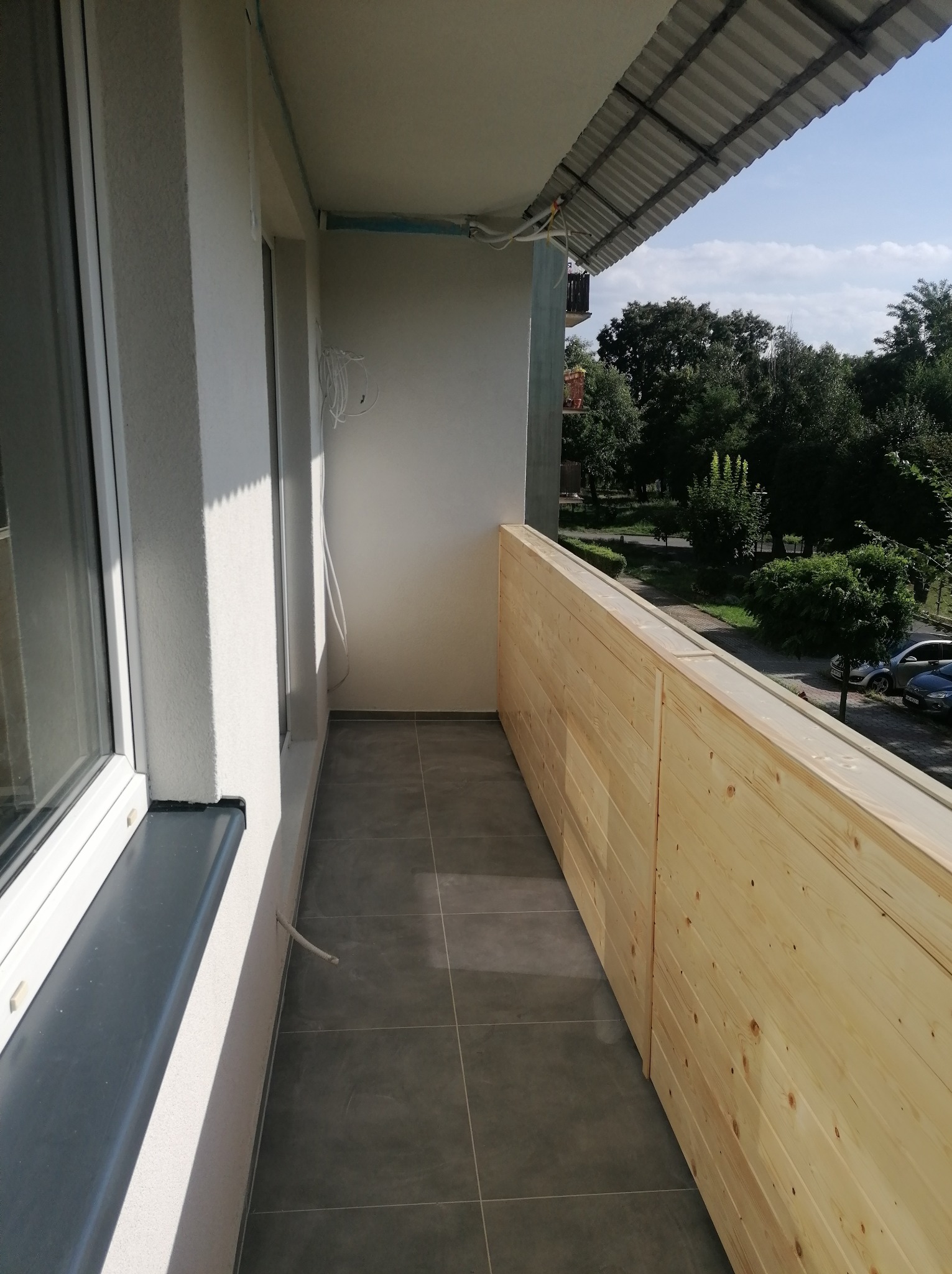 Wykończony balkon z szarą podłogą z płytek i nową drewnianą balustradą z jasnego drewna, widok na zieleń i drzewa.