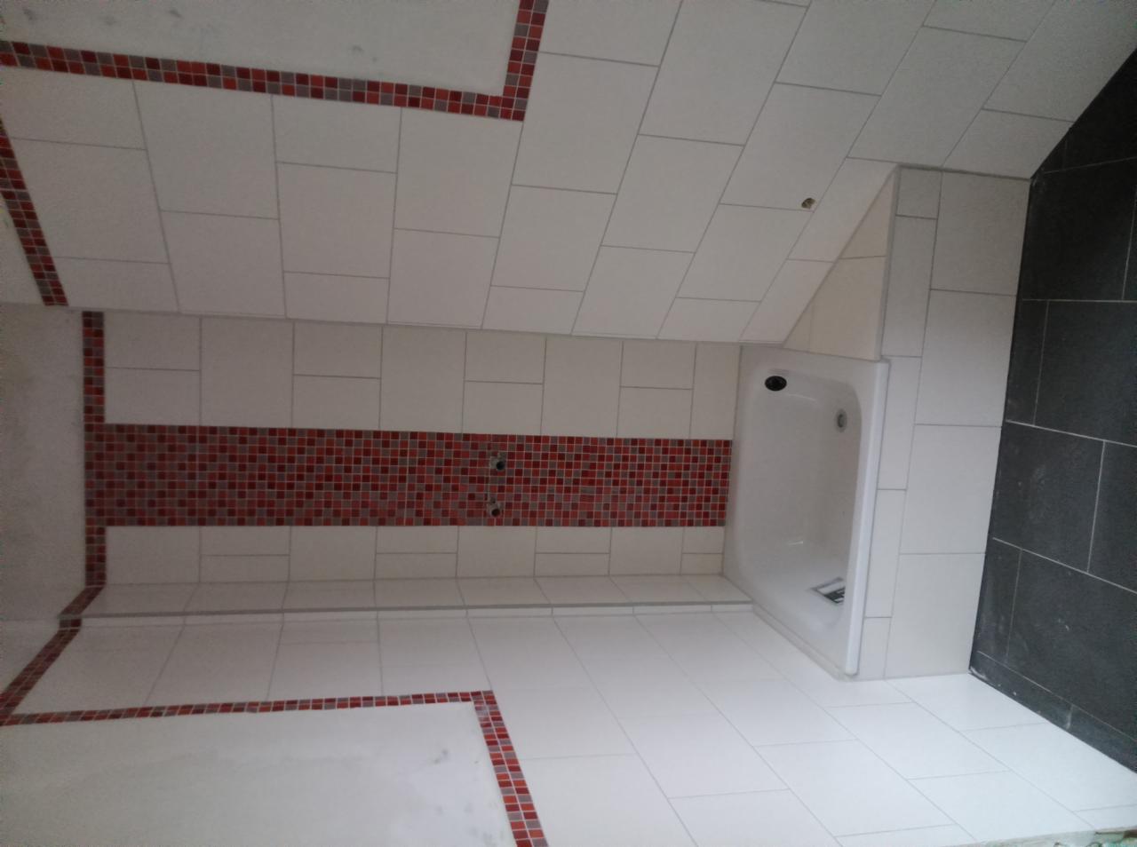 Wykończona łazienka na poddaszu z białą wanną, białymi płytkami i dekoracyjnym pasem czerwonej mozaiki. Ciemnoszara ściana z płytek po prawej stronie.