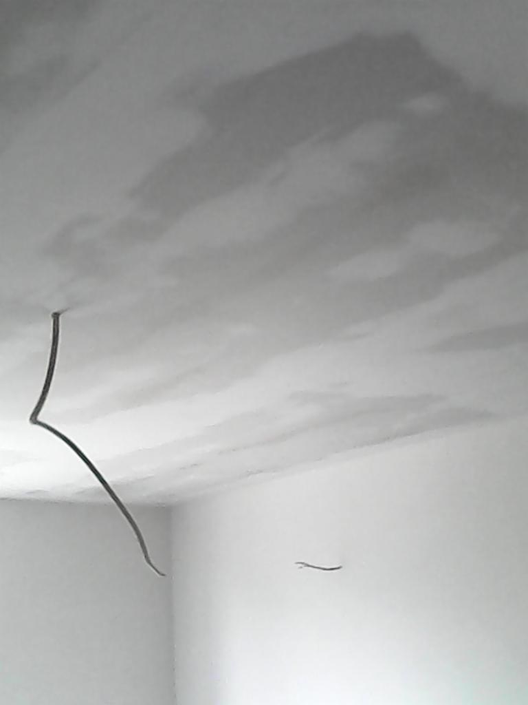 Surowy stan wnętrza: Świeżo nałożona, jeszcze wilgotna gładź na suficie, widoczne niedoskonałości i wystające przewody elektryczne, białe ściany w narożniku pokoju.