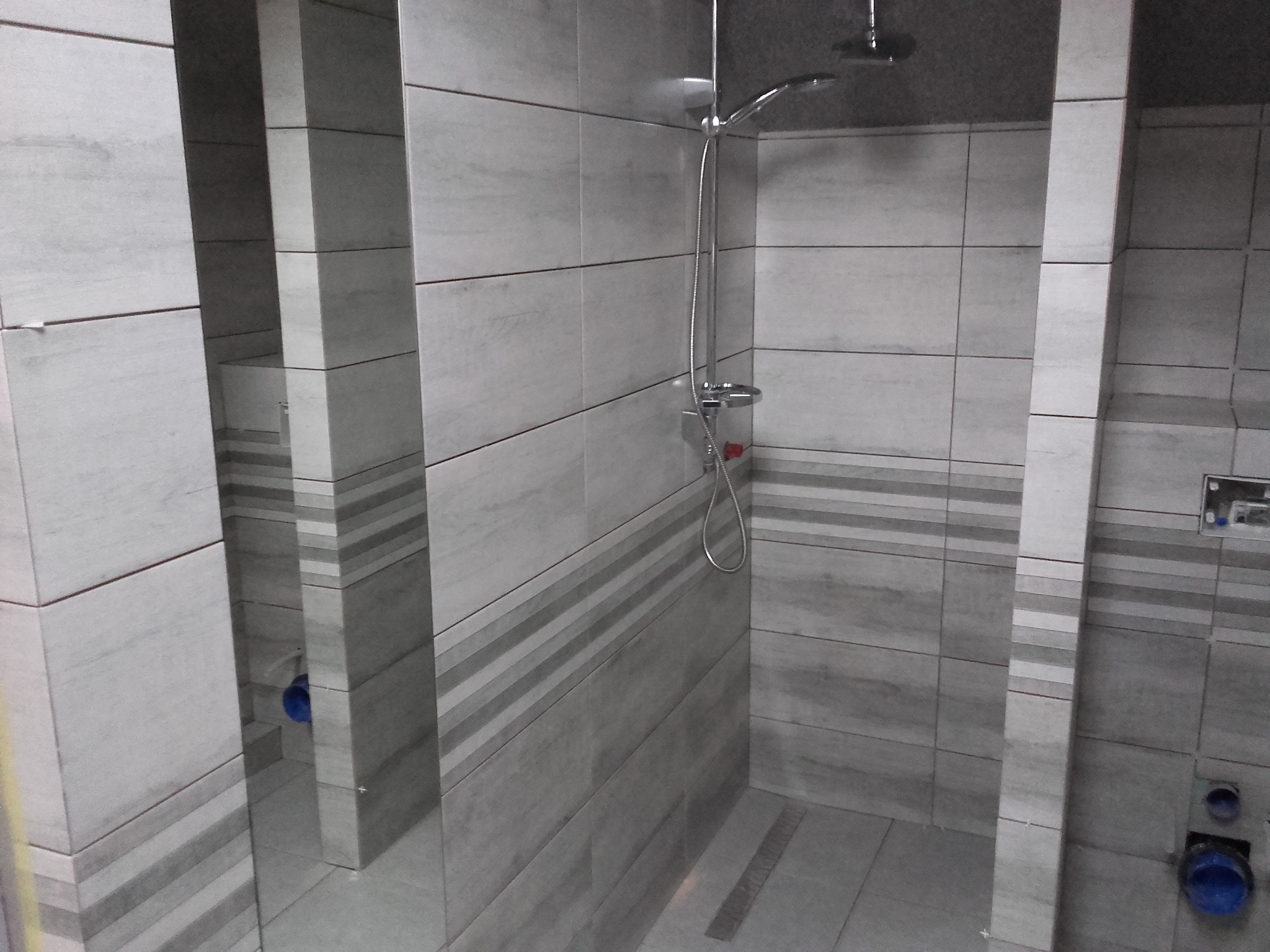 Szara kabina prysznicowa wyłożona płytkami z poziomymi paskami, widoczny odpływ liniowy w podłodze i bateria prysznicowa.