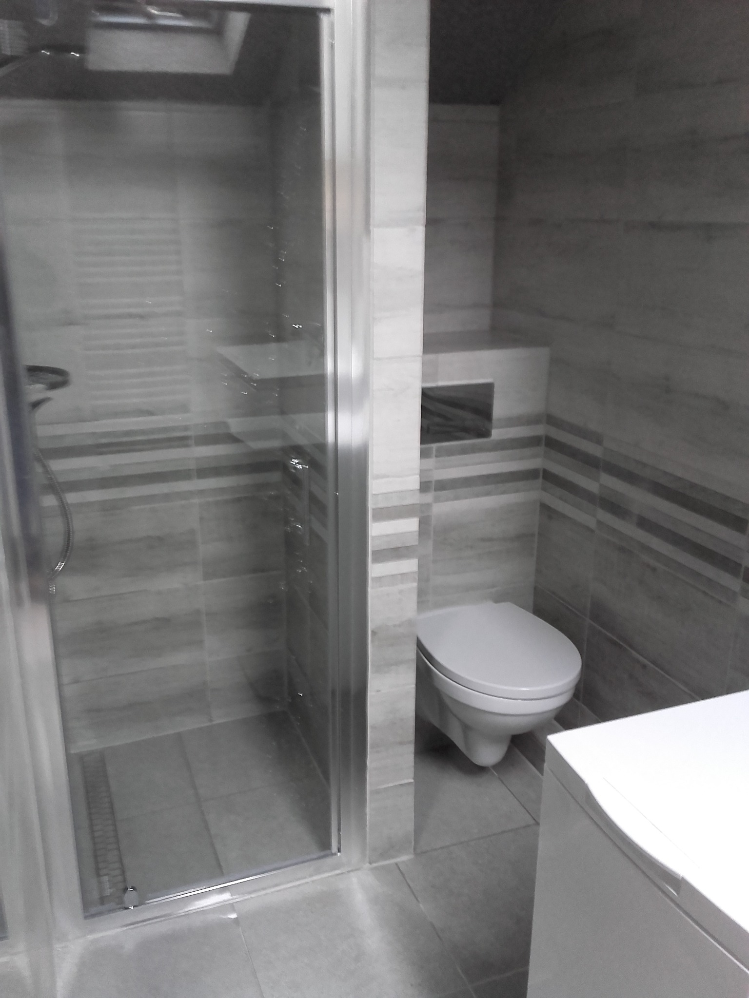 Szaro-biała łazienka po remoncie: kabina prysznicowa ze szklanymi drzwiami, wisząca toaleta z ukrytym spłukiwaniem, płytki imitujące drewno z poziomymi pasami, biała szafka pod umywalkę.