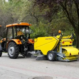 Żółty traktor z zamiatarką Brodd Twiggy czyszczący kostkę brukową w parku. Widoczne drzewa i zieleń w tle, co sugeruje sprzątanie w przestrzeni miejskiej.