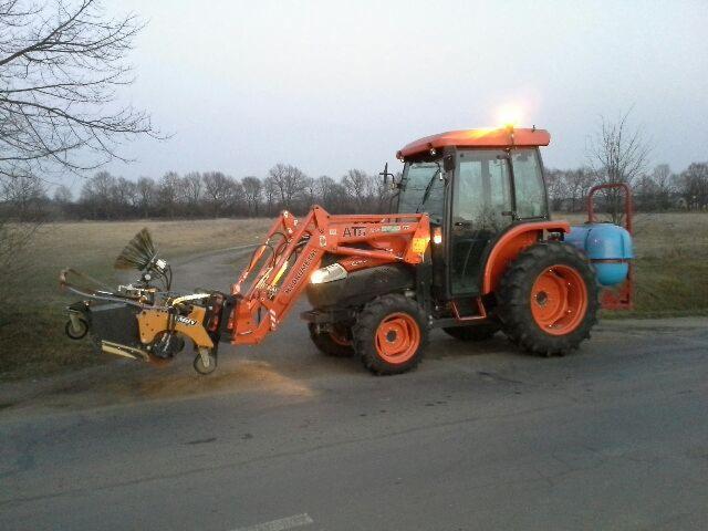 Pomarańczowy traktor z zamontowaną szczotką na drodze asfaltowej, gotowy do zamiatania. Widoczne drzewa w tle, pochmurne niebo.