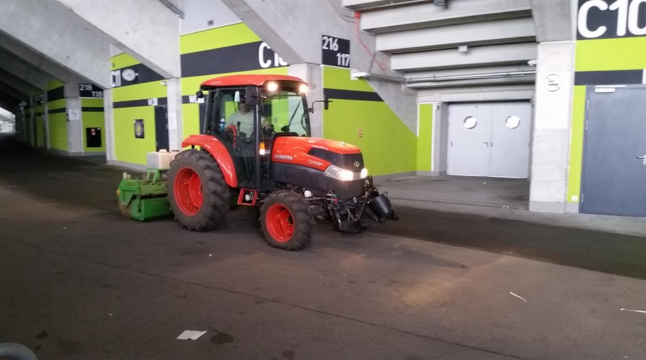 Pomarańczowy traktor Kubota z zielonym osprzętem czyszczącym, na tle betonowego parkingu hali z numeracją sektorów C10, 216, 218. Widoczne drzwi wejściowe.