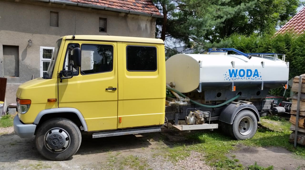 Żółta cysterna z napisem 'WODA' zaparkowana na trawie przed budynkiem. Widoczne detale pojazdu i otoczenie wiejskie.