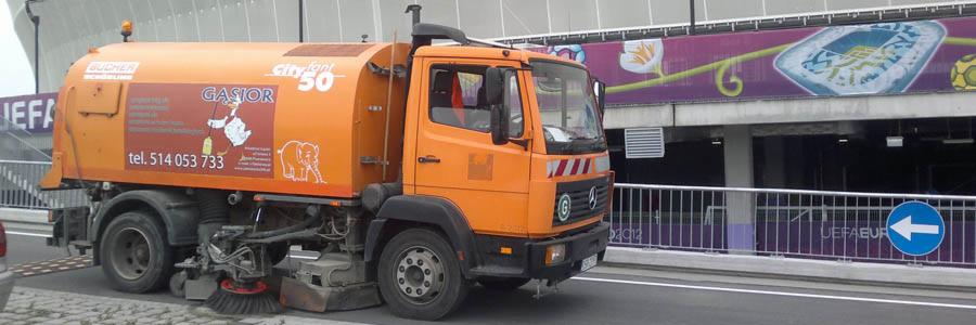 Pomarańczowa zamiatarka uliczna marki Bucher z logo firmy Gasior na tle stadionu i znaku drogowego. Widoczne szczotki czyszczące jezdnię.