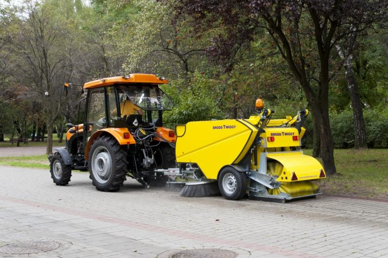 Żółty traktor z zamiatarką Brodd Twiggy czyszczący kostkę brukową w parku. Widoczne drzewa i zieleń w tle, co sugeruje sprzątanie w przestrzeni miejskiej.